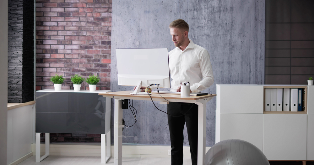 A professional man standing at a sleek standing desk in a modern office setting, with visible wire management under the desk. The office features a clean design with potted plants, brick and textured walls, and organised storage units.