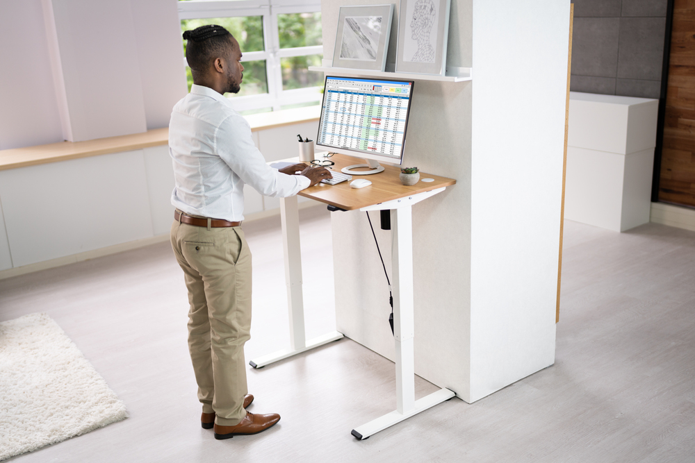 A man working on a computer at a modern standing desk in a bright and well-lit office space. The workspace features organised desk accessories and a natural, minimalist design.